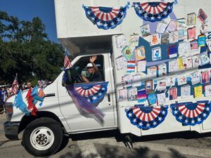 A librarian waving out of the window of the Bookmobile, which is decorated in colorful cut outs of books and 4th of July memorabilia.