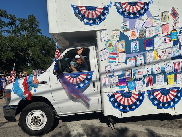 A librarian waving out of the window of the Bookmobile, which is decorated in colorful cut outs of books and 4th of July memorabilia.